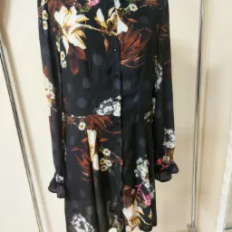 Czarna, zapinana na guziki sukienka z długim rękawem, w kwiatowy wzór, na manekinie, prezentowana na tle kremowej ściany i chromowanego wieszaka.