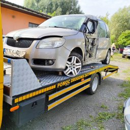 TOM-TRANS POMOC DROGOWA 24/7 - Uszkodzony samochód marki Skoda z powybijanymi szybami, brakiem drzwi i uszkodzeniami karoserii przewożony na żółtej lawecie z napisem 'POMOC DROGOWA 24/7'.