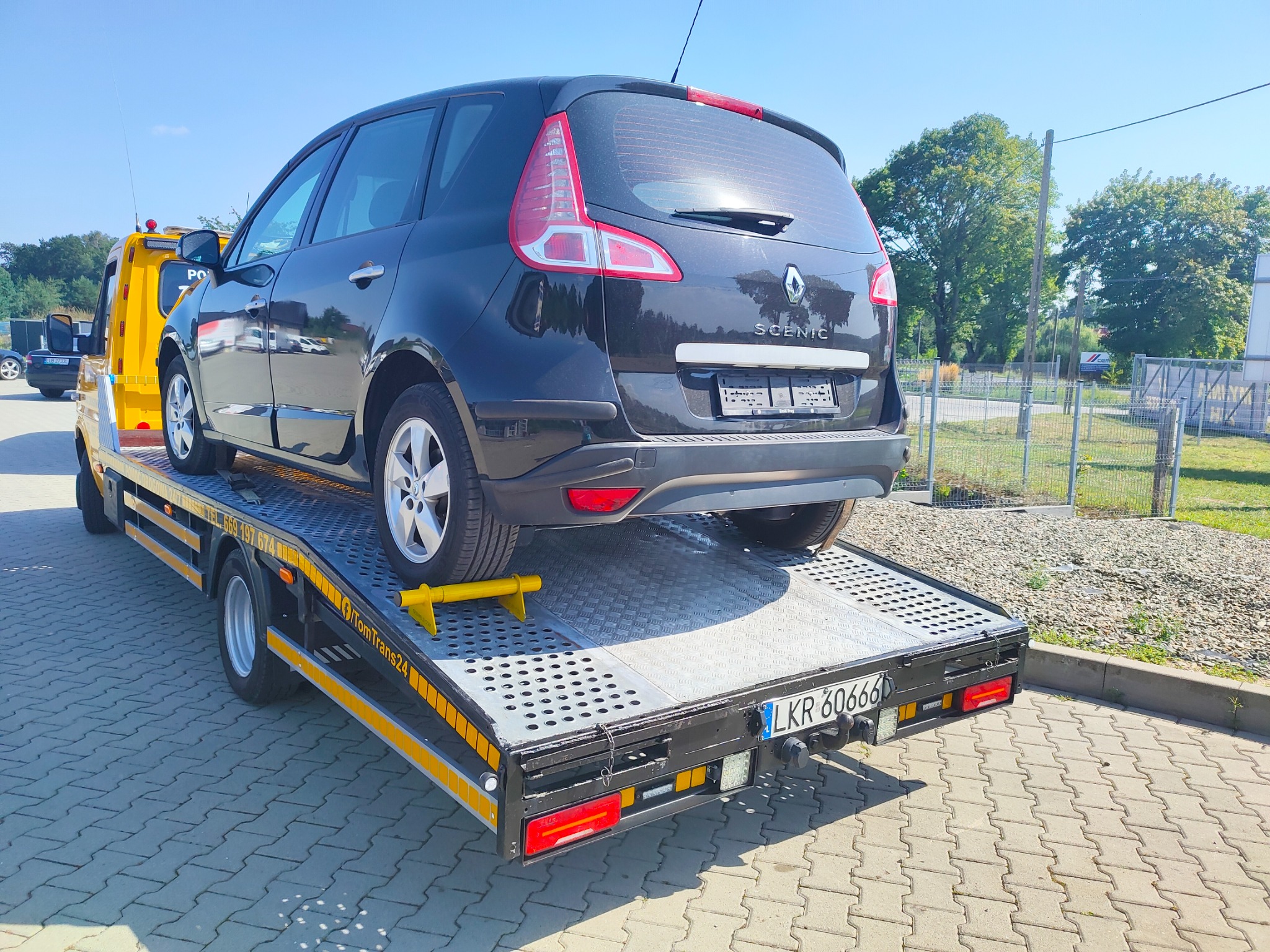 Czarny Renault Scenic zabezpieczony żółtą blokadą na lawecie z widoczną tablicą rejestracyjną LKR 60666, w tle zieleń i błękitne niebo.