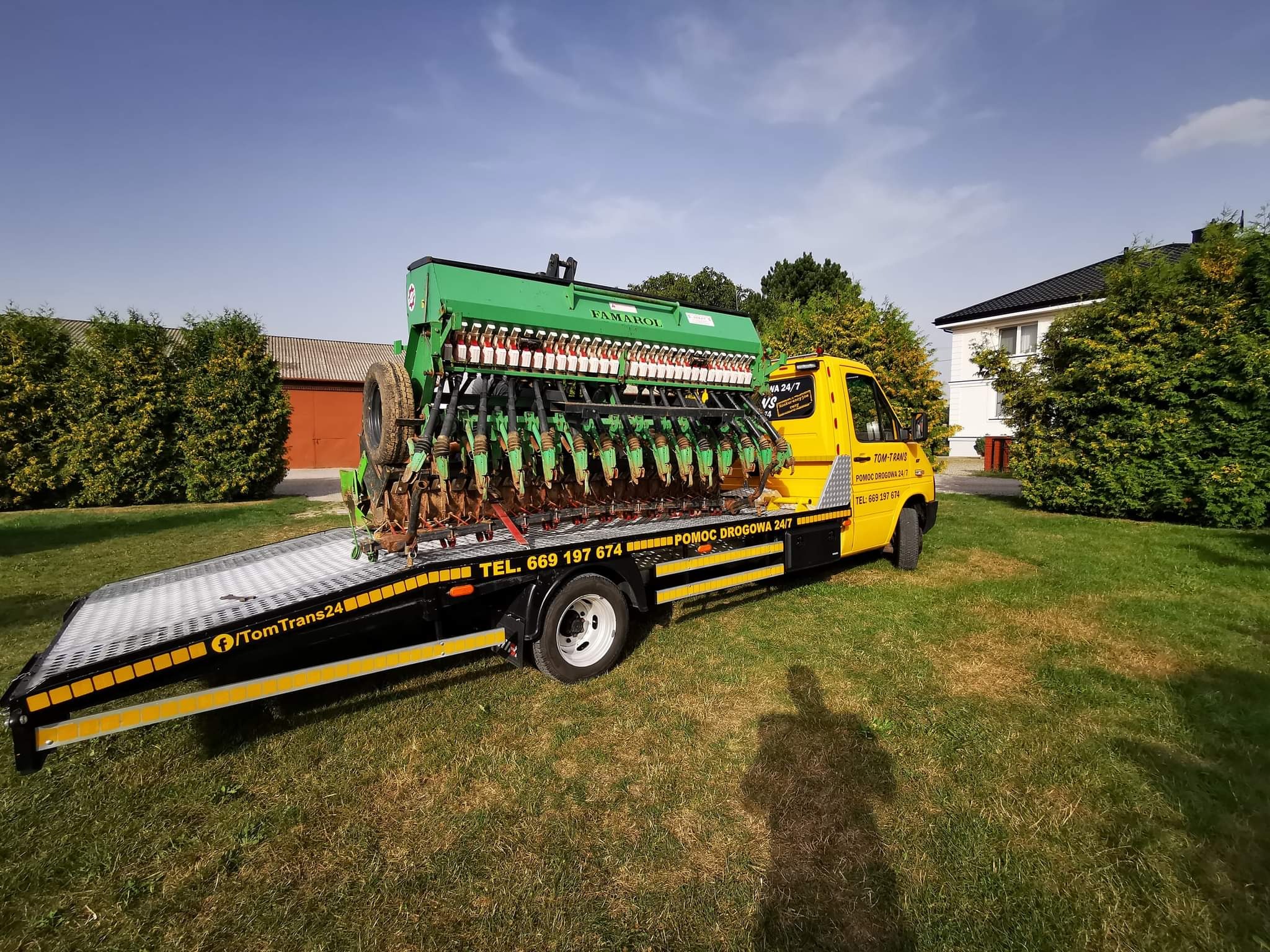 Żółta laweta TomTrans24 z Kraśnika przewozi zielony siewnik rolniczy marki FAMAROL na tle budynku i trawnika.