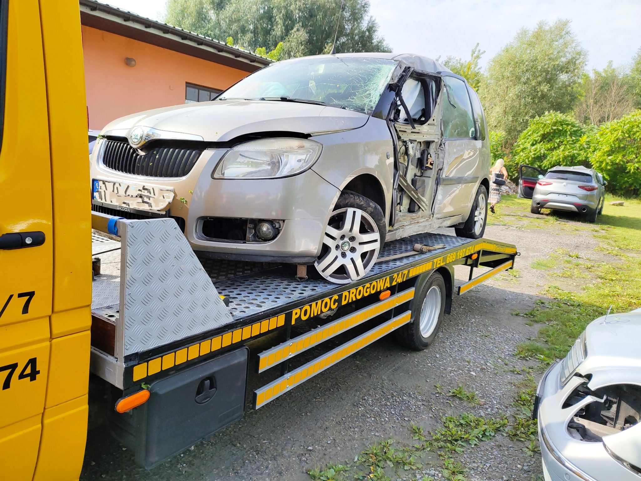 Uszkodzony samochód marki Skoda z powybijanymi szybami, brakiem drzwi i uszkodzeniami karoserii przewożony na żółtej lawecie z napisem 'POMOC DROGOWA 24/7'.