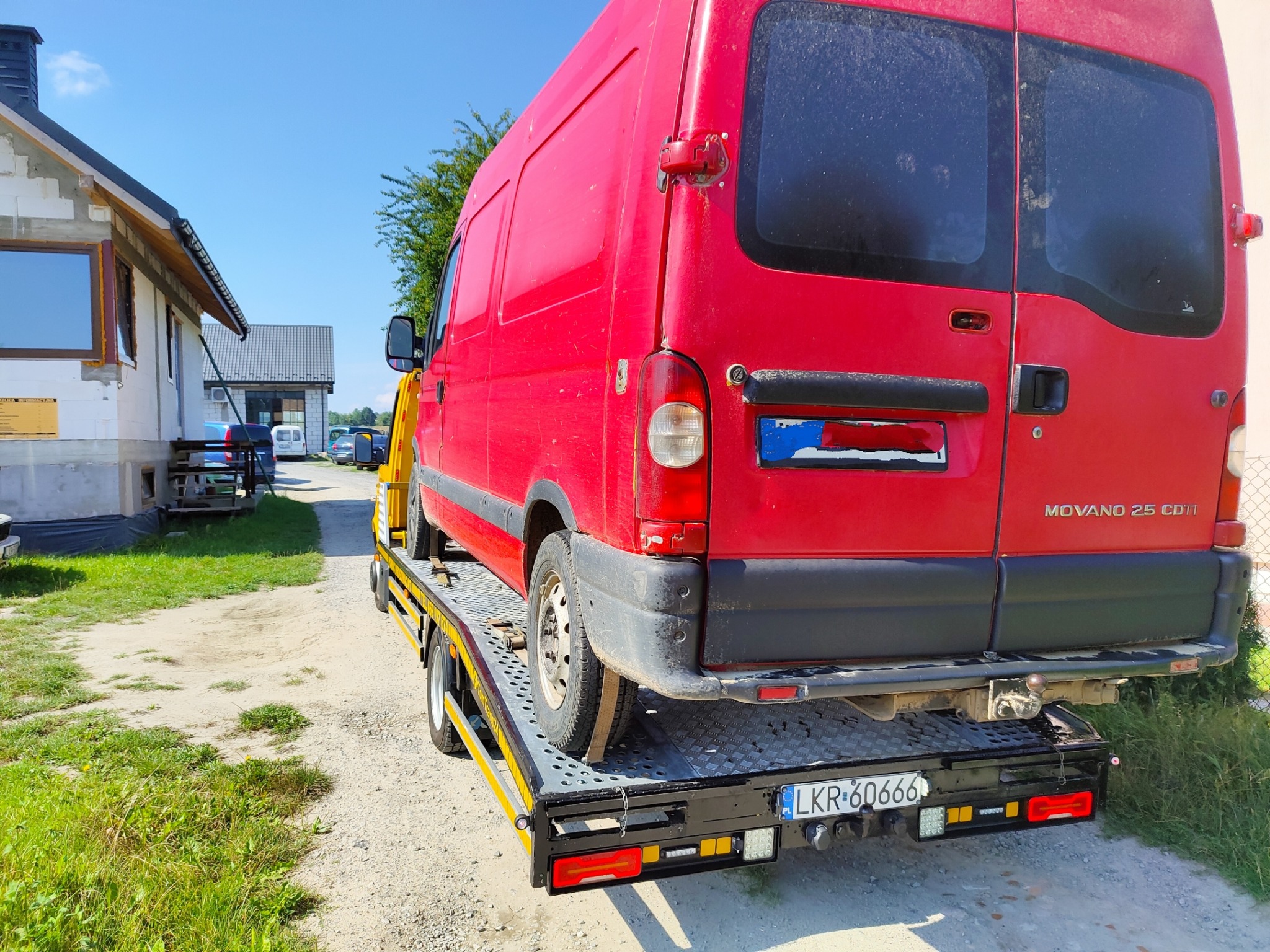 Czerwony bus marki Movano 2.5 CDTI transportowany na żółto-czarnej lawecie na tle budowy domu.