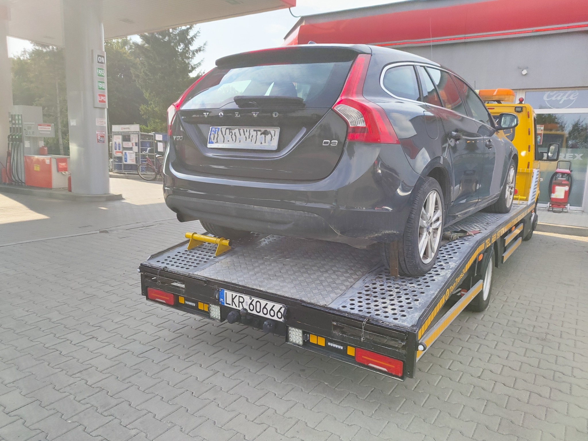 Czarny samochód Volvo V60 na platformie lawety na tle stacji benzynowej w słoneczny dzień. Widoczne zabezpieczenia kół.