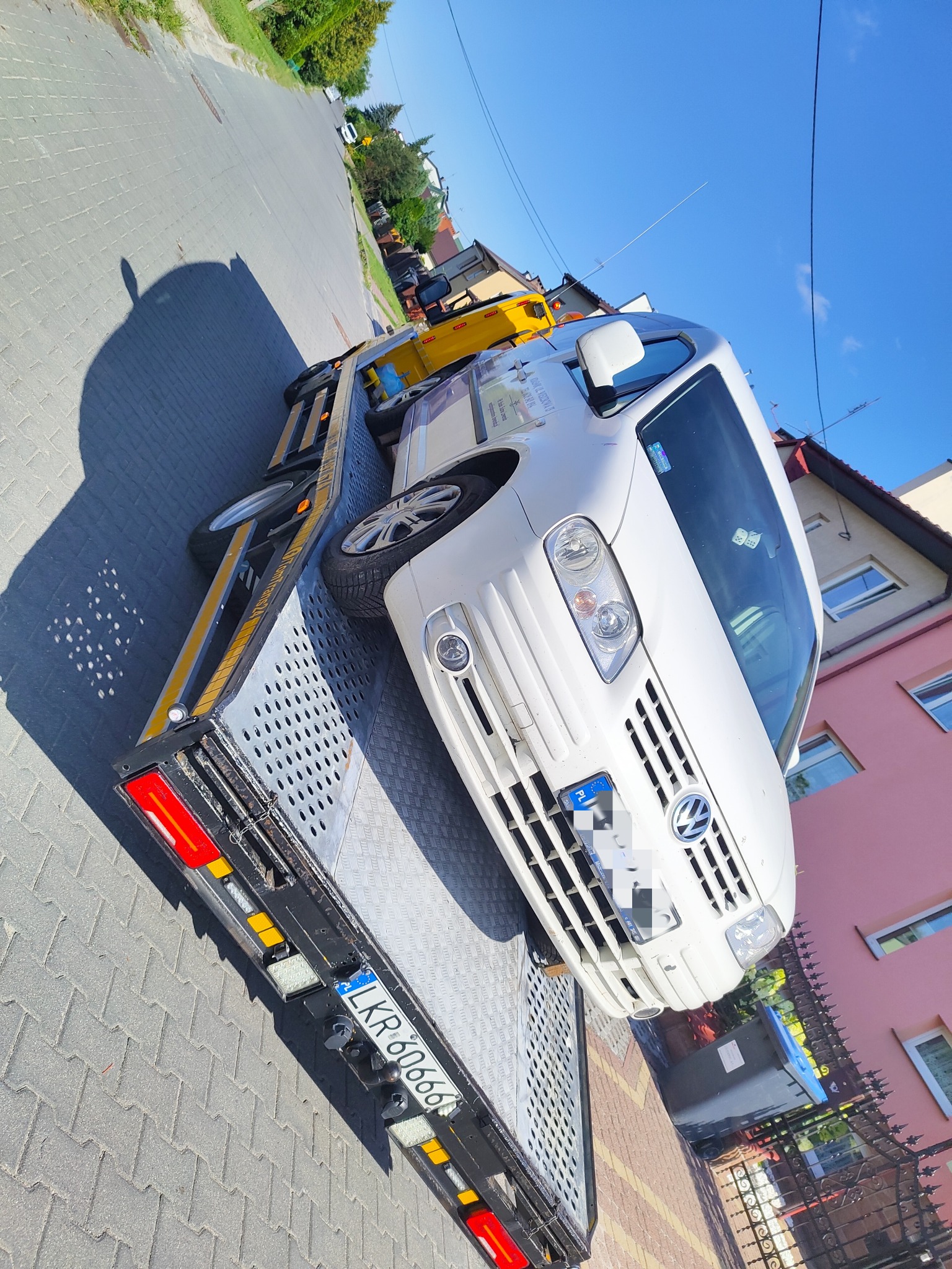 Biały Volkswagen Caddy na platformie lawety transportowany ulicą w słoneczny dzień. Widoczny numer rejestracyjny lawety LKR 60666.