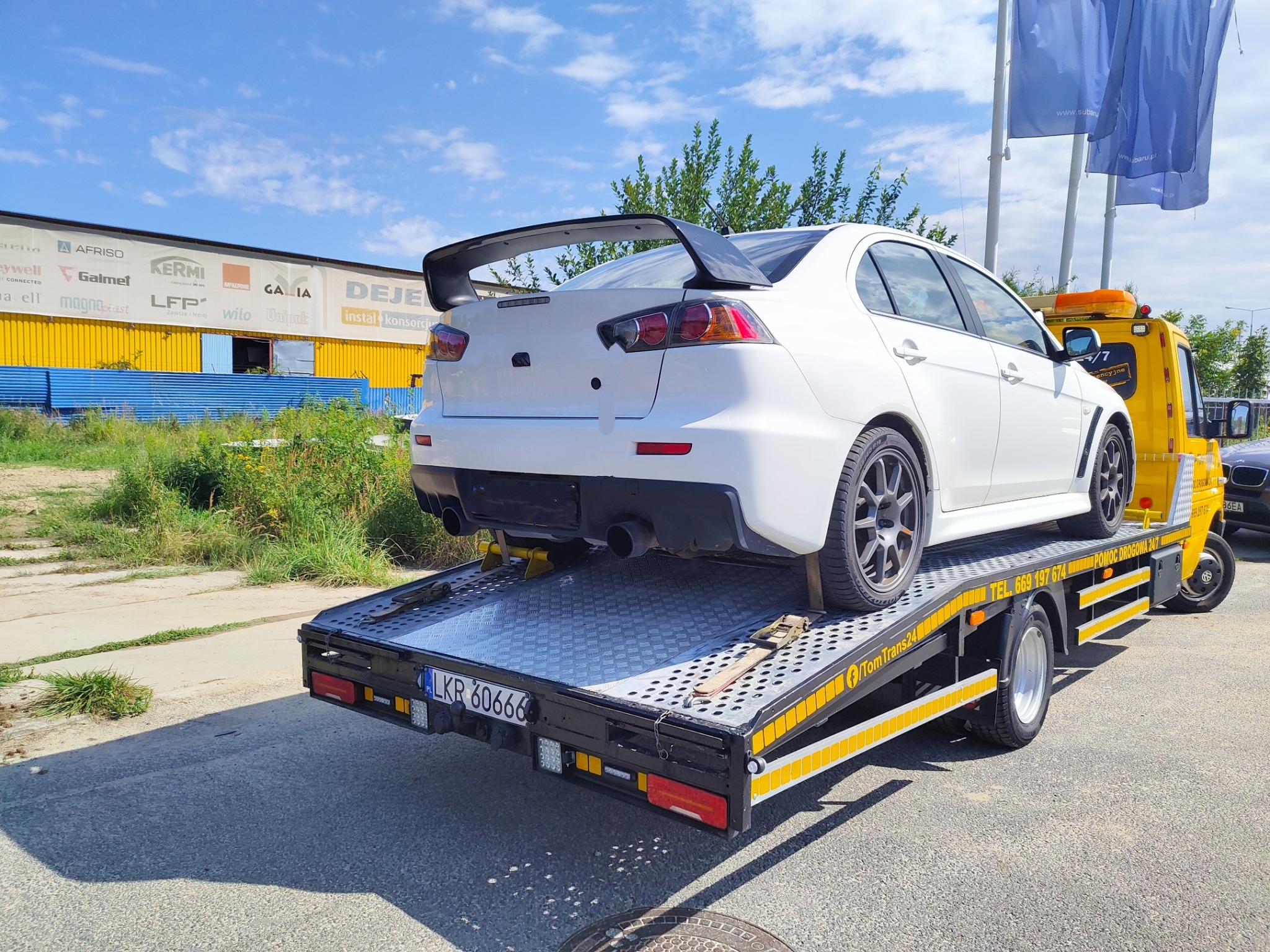 Biały sportowy samochód z dużym spoilerem załadowany na żółtą lawetę pomocy drogowej z numerem rejestracyjnym LKR 60666, w tle budynek z reklamami firm.