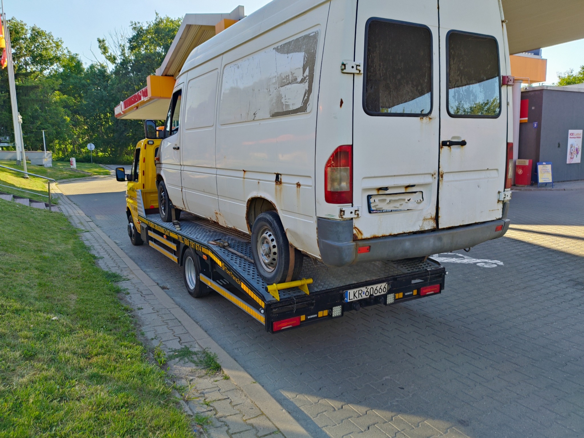Żółta laweta z białym, lekko zardzewiałym busem na platformie, stojąca na kostce brukowej w pobliżu stacji benzynowej w słoneczny dzień.