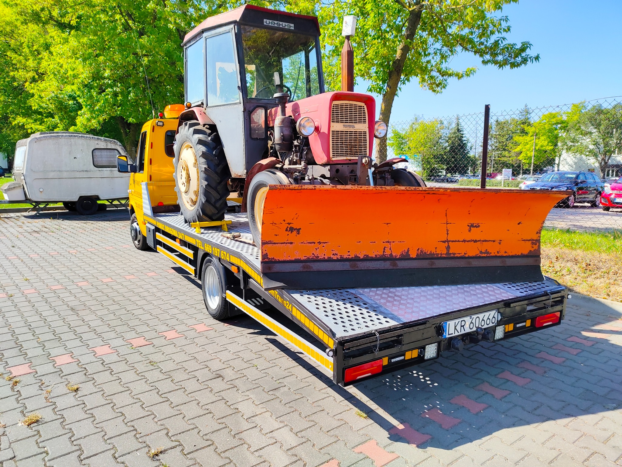 Czerwony traktor Ursus z pomarańczowym pługiem na żółtej lawecie z tablicą rejestracyjną LKR 60666, obok przyczepa kempingowa, na tle zieleni i błękitnego nieba.