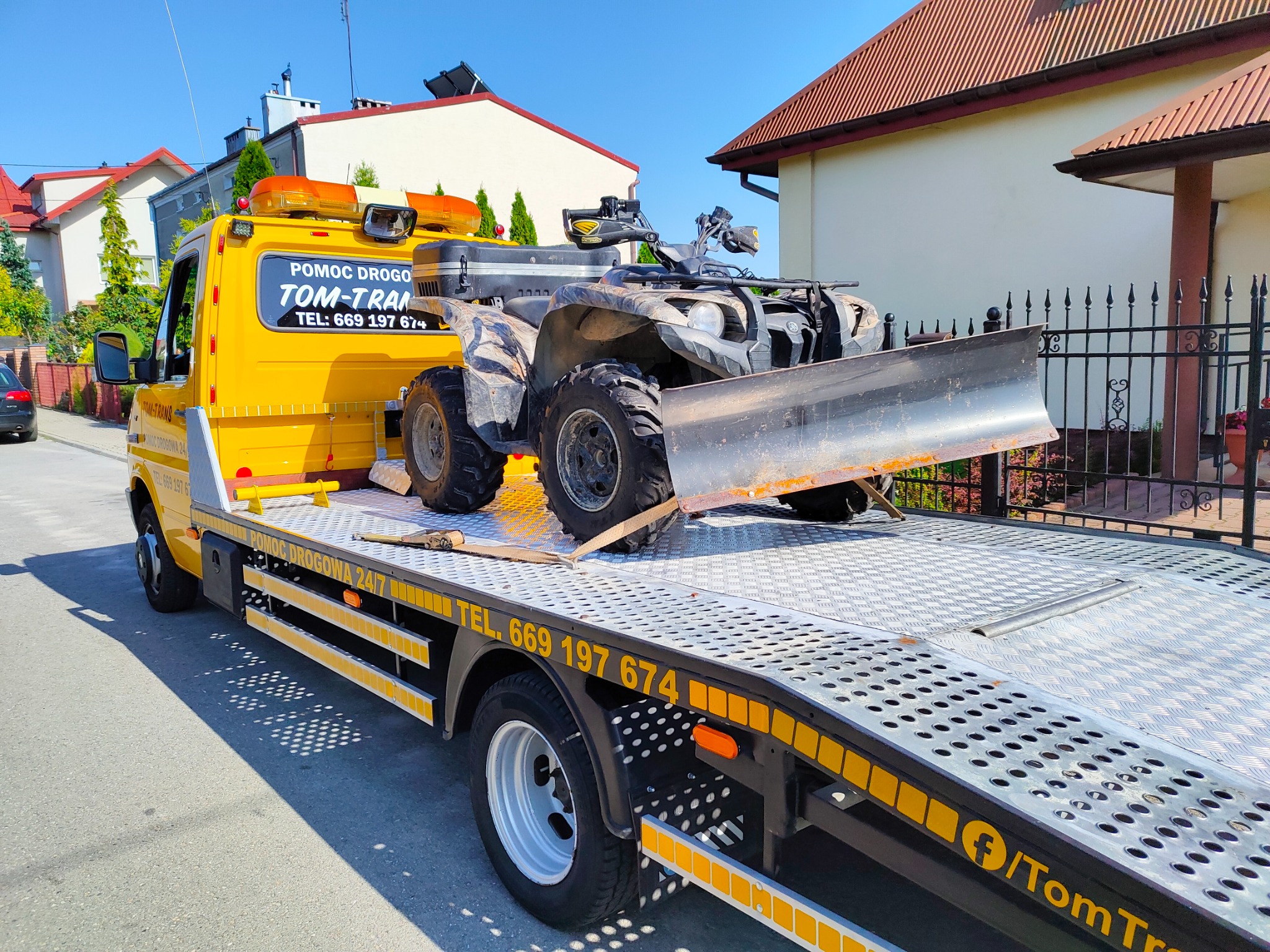 Żółta laweta z napisem 'Pomoc Drogowa' i numerem telefonu przewozi brudnego quada z pługiem na tle zabudowań mieszkalnych.