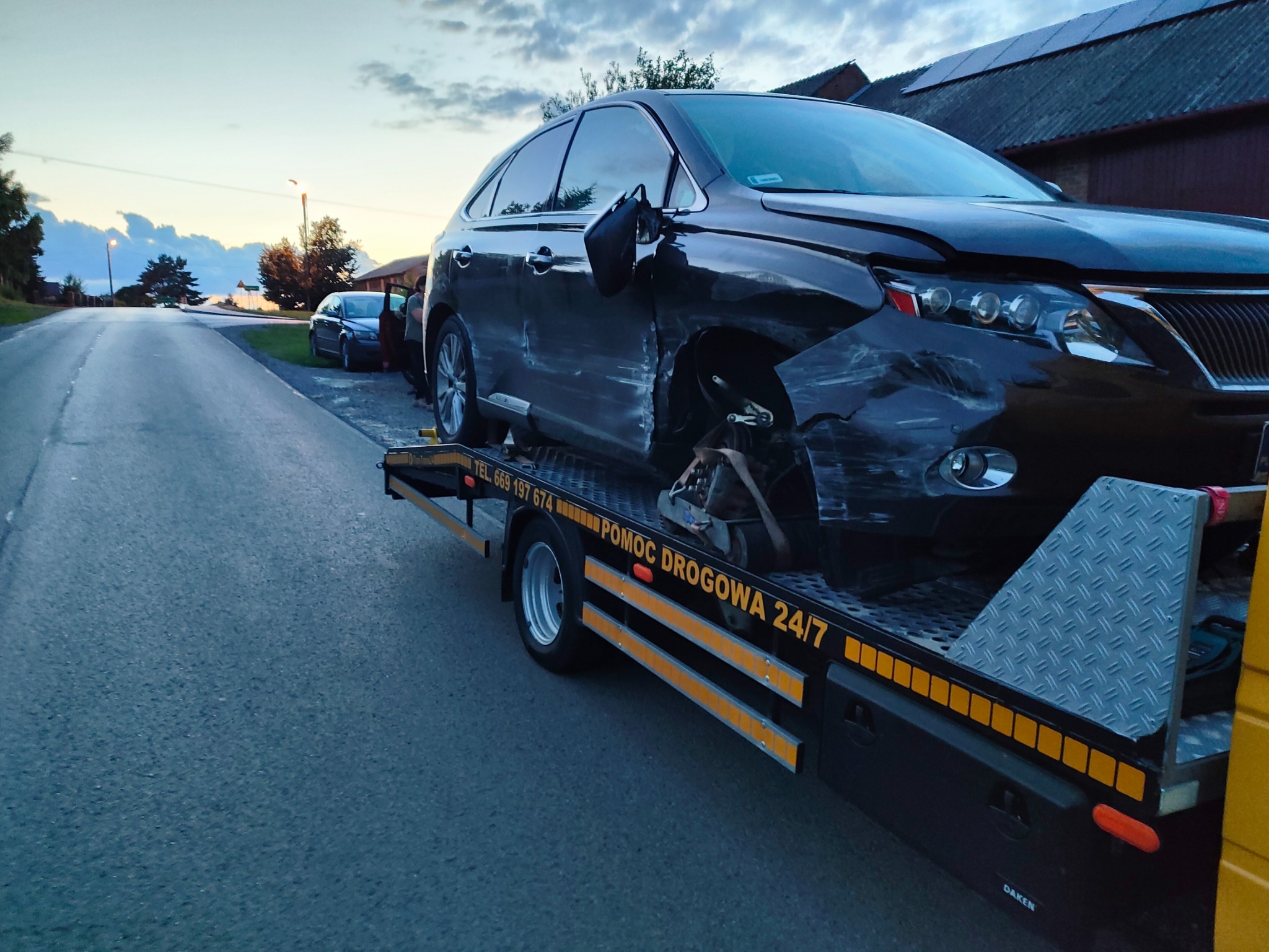 Czarny uszkodzony samochód osobowy marki Lexus załadowany na platformę lawety pomocy drogowej na tle wiejskiej drogi i zabudowań gospodarczych