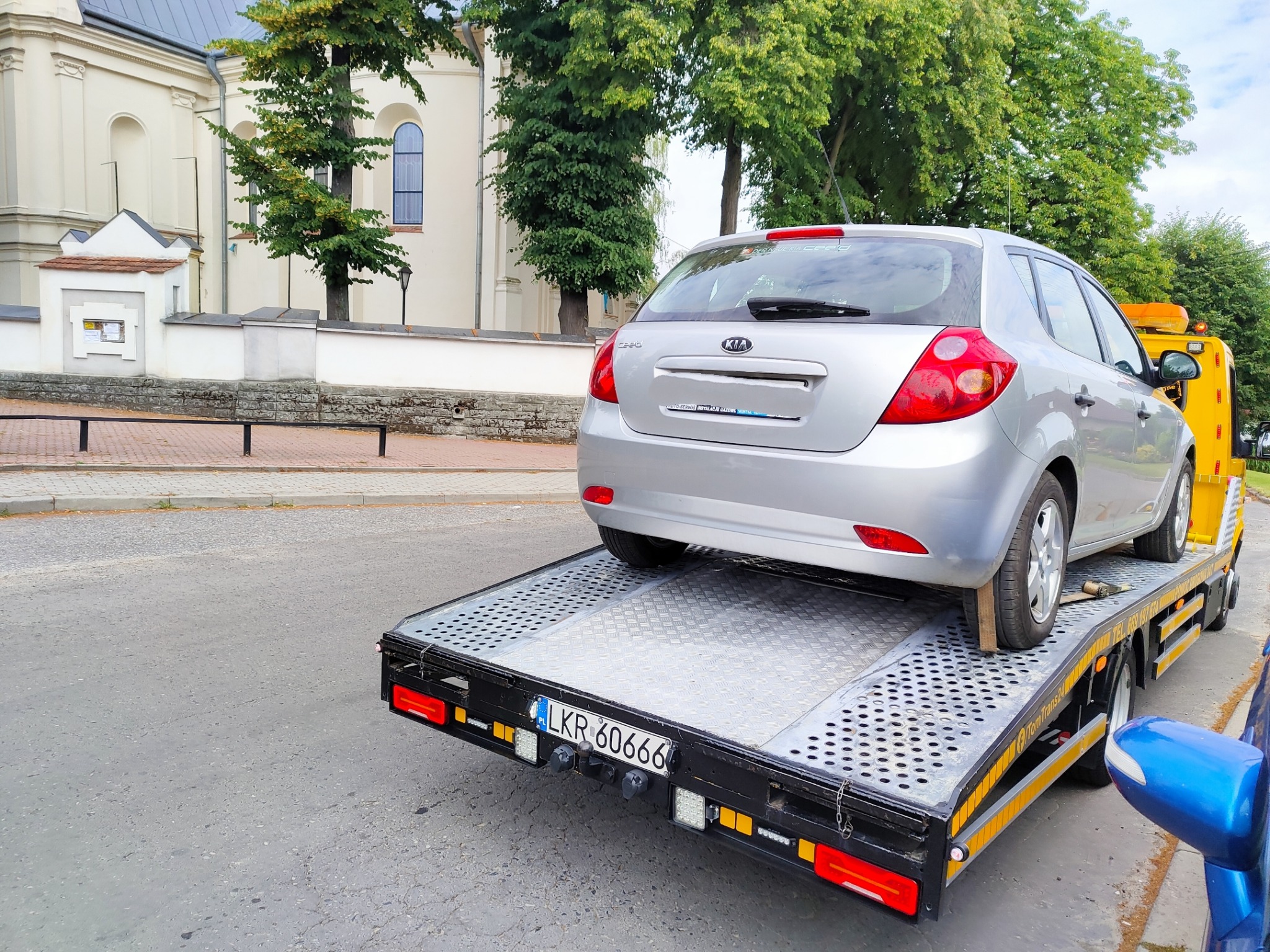 Srebrny samochód Kia Ceed na platformie lawety z widoczną tablicą rejestracyjną LKR 60666, w tle budynek kościoła i drzewa.