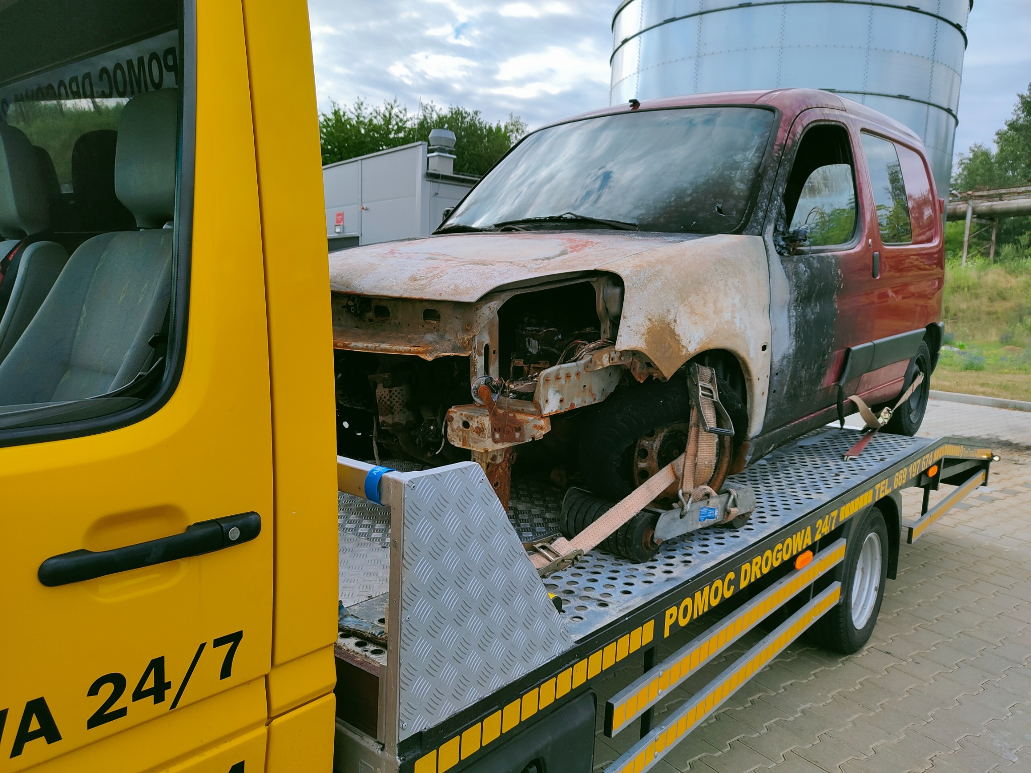 Uszkodzony, spalony samochód osobowy zabezpieczony pasami transportowymi na platformie lawety pomocy drogowej z napisem 'POMOC DROGOWA 24/7'. W tle widoczny silos i zieleń.