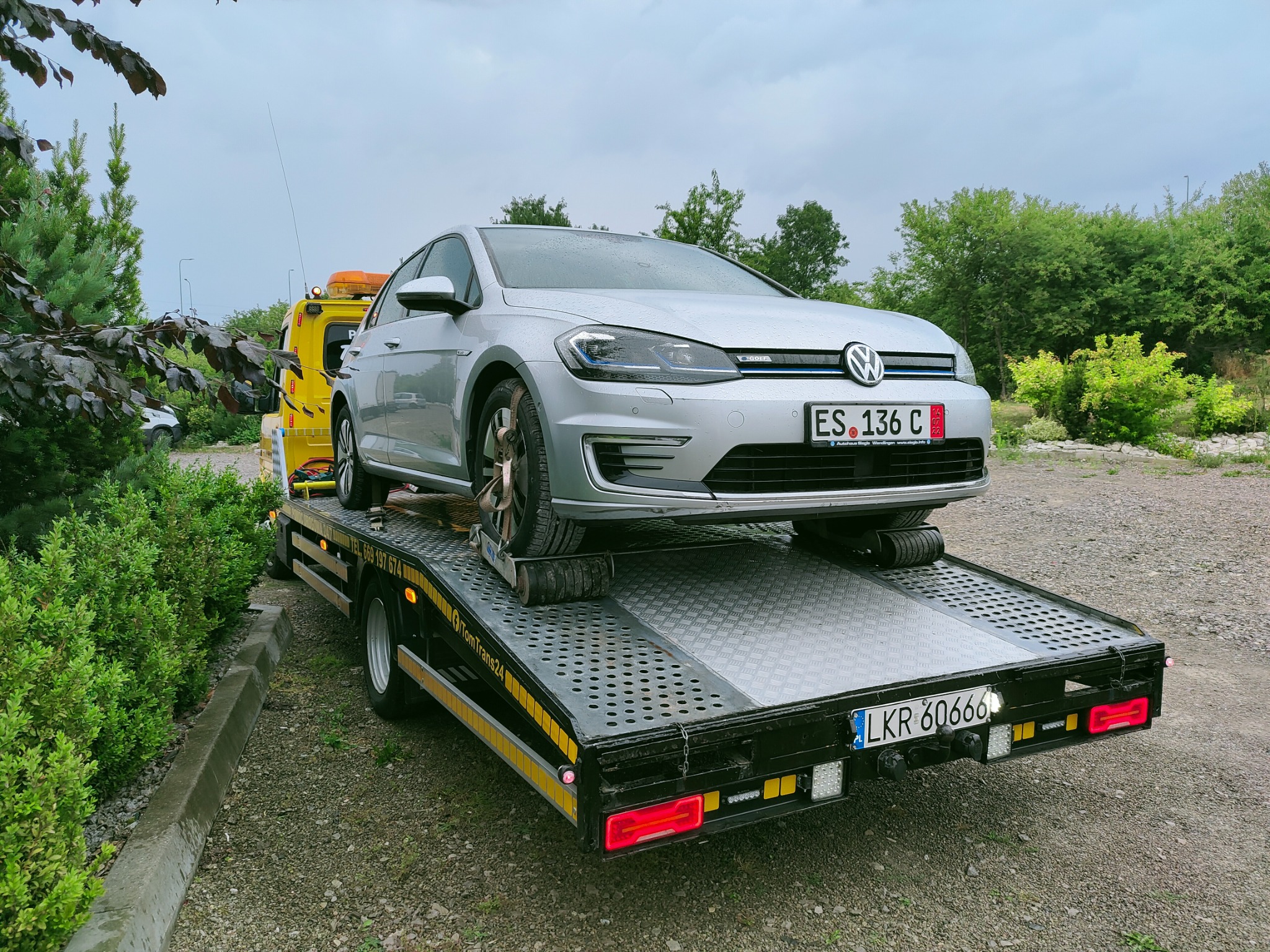Srebrny Volkswagen e-Golf z hiszpańską tablicą rejestracyjną ES-136-C umieszczony na czarnej lawecie TomTrans24 z polską tablicą LKR 60666, sfotografowany w pochmurny dzień na żwirowym podłożu.