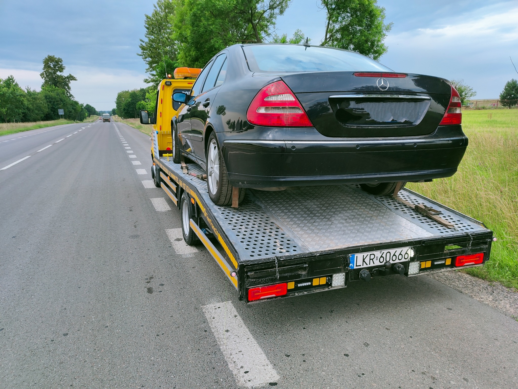 Czarny samochód osobowy marki Mercedes-Benz zabezpieczony na żółtej lawecie, widok z tyłu, tablica rejestracyjna LKR-60666, droga asfaltowa z poboczem w tle.