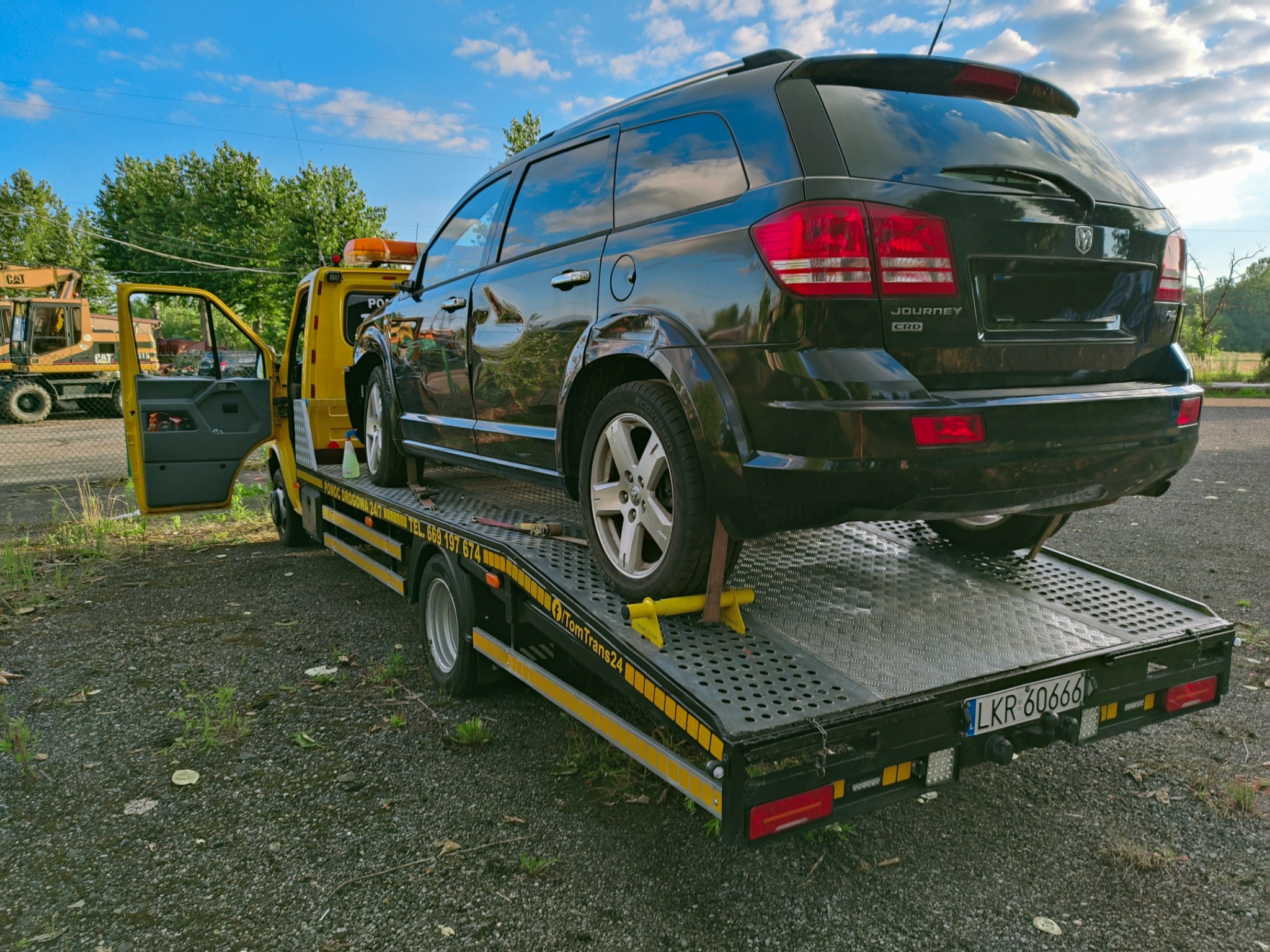 Czarny samochód Dodge Journey CRD zabezpieczony żółtymi blokadami na platformie lawety pomocy drogowej, z otwartymi drzwiami kierowcy i widocznym numerem rejestracyjnym lawety LKR 60666, w tle...