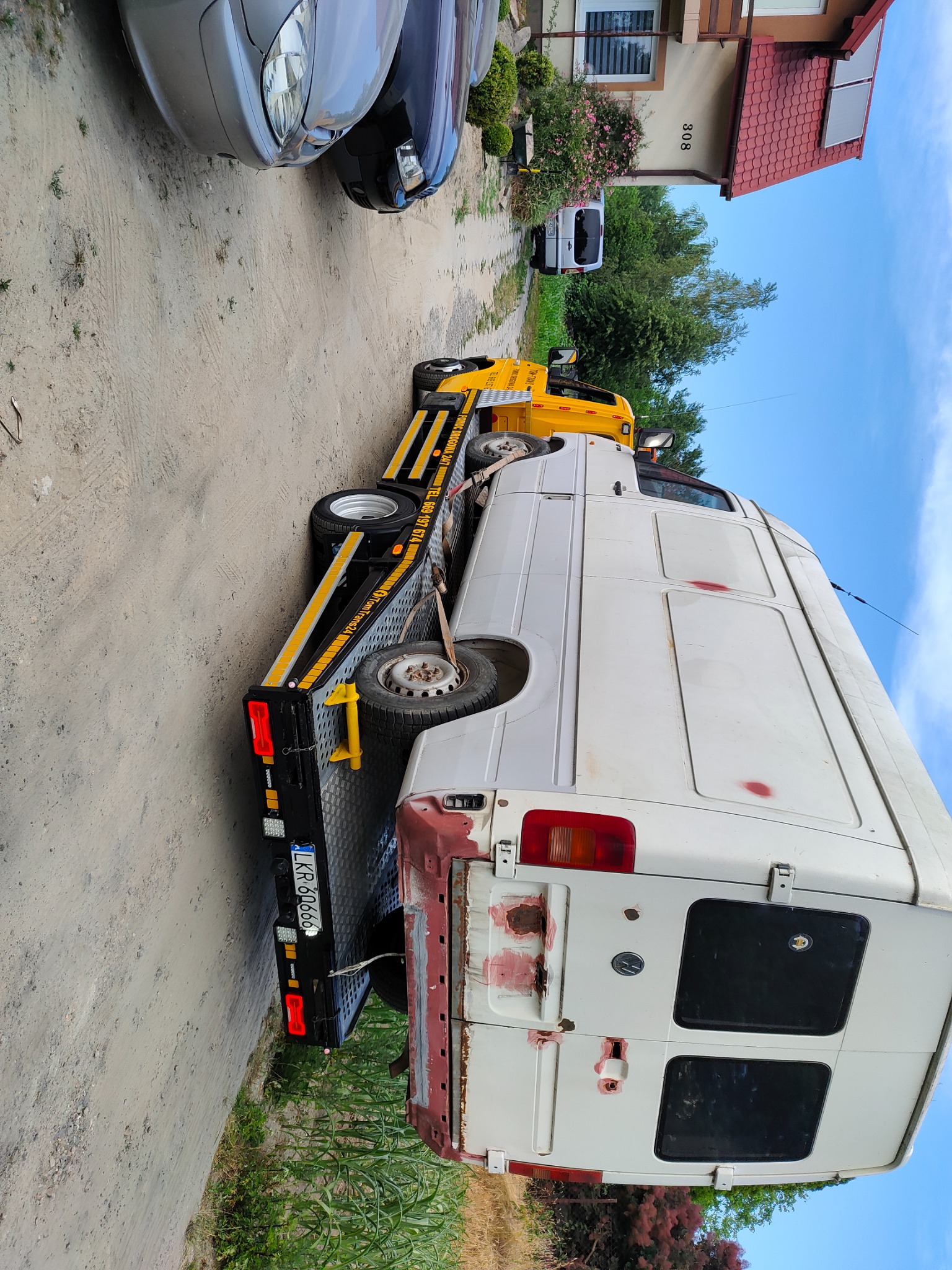 Uszkodzony biały van na żółto-czarnej lawecie z widocznymi śladami rdzy i napraw blacharskich, zaparkowany na żwirowej drodze obok domu z czerwonym dachem.