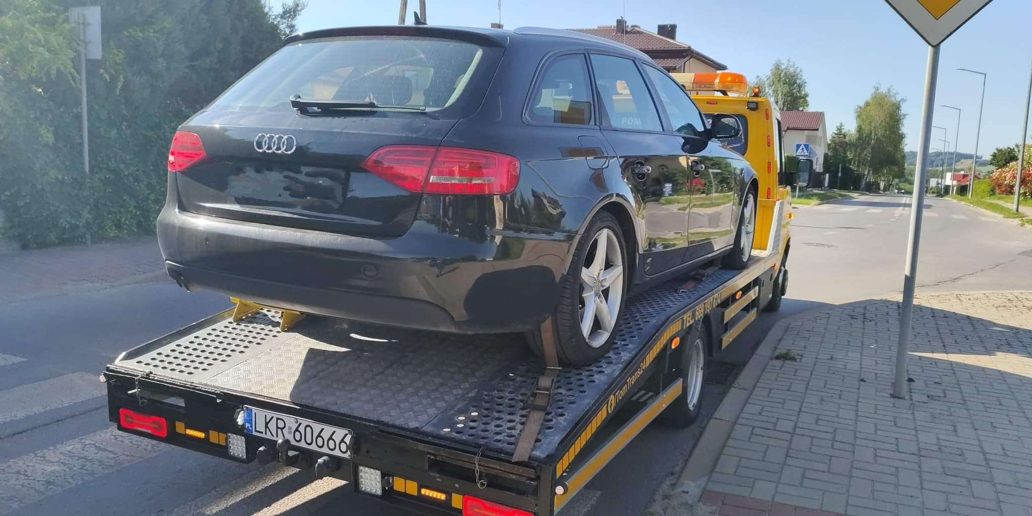 Czarny samochód marki Audi kombi z numerem rejestracyjnym LKR 60666, zabezpieczony pasami, na platformie żółtej lawety z włączonymi światłami ostrzegawczymi na tle ulicy i zabudowań.