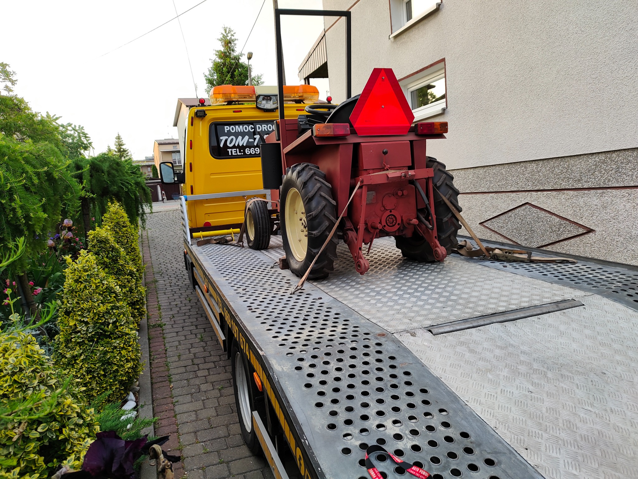 Żółta laweta z czerwonym traktorem rolniczym na platformie, widok z tyłu, z widocznym numerem telefonu pomocy drogowej na kabinie