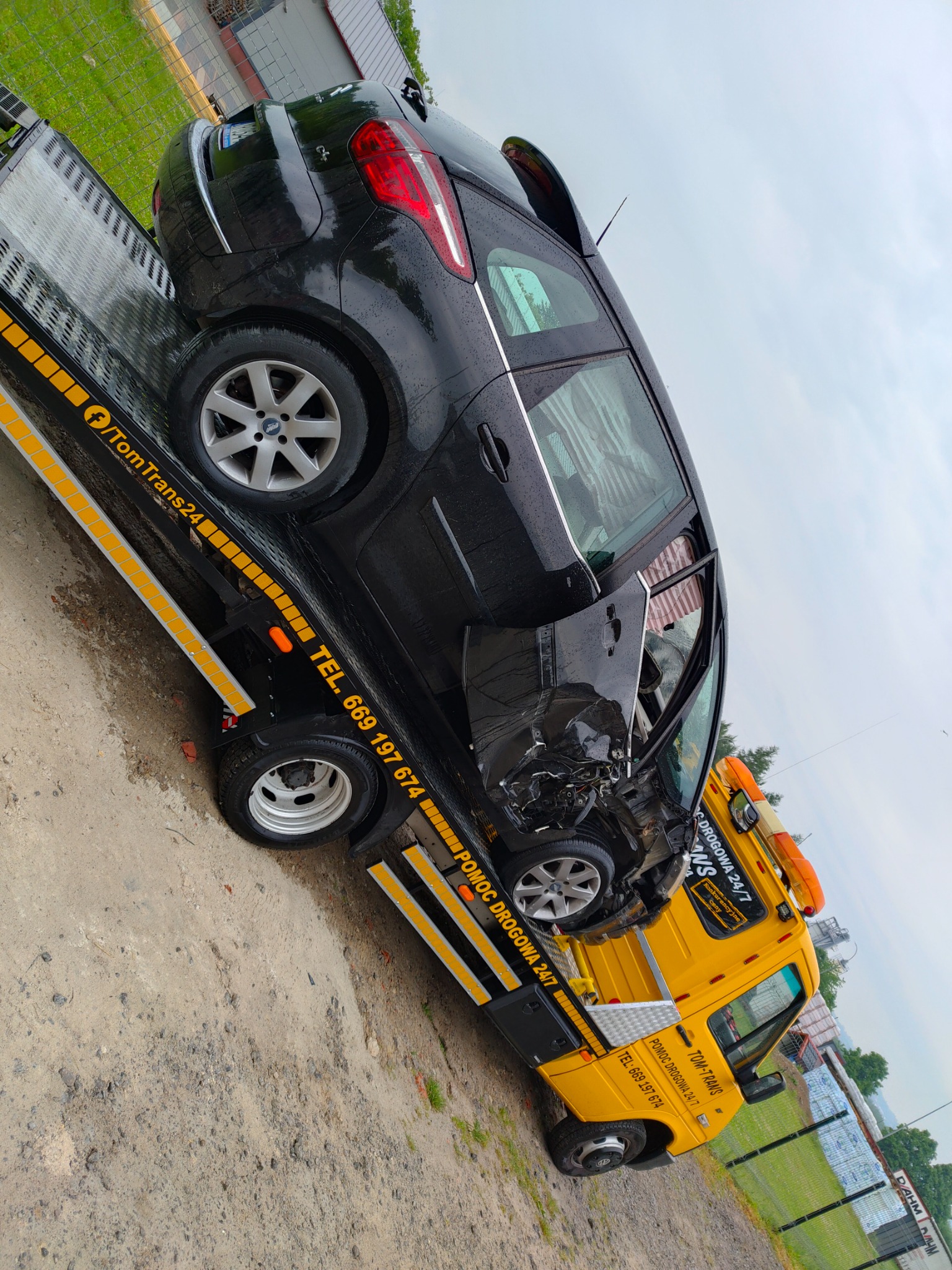 Czarny uszkodzony samochód osobowy na żółtej lawecie z widocznym numerem telefonu pomocy drogowej i logo firmy TomTrans24.