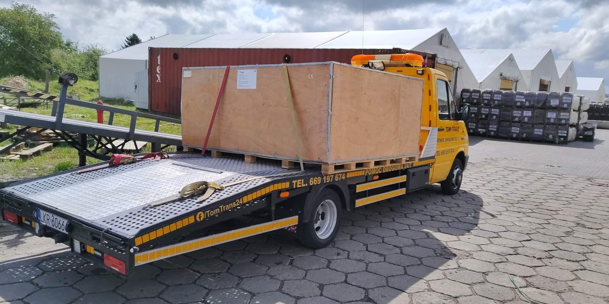 Żółta laweta z drewnianą skrzynią na palecie, zabezpieczoną pasami transportowymi, na tle magazynowych namiotów i kontenera.