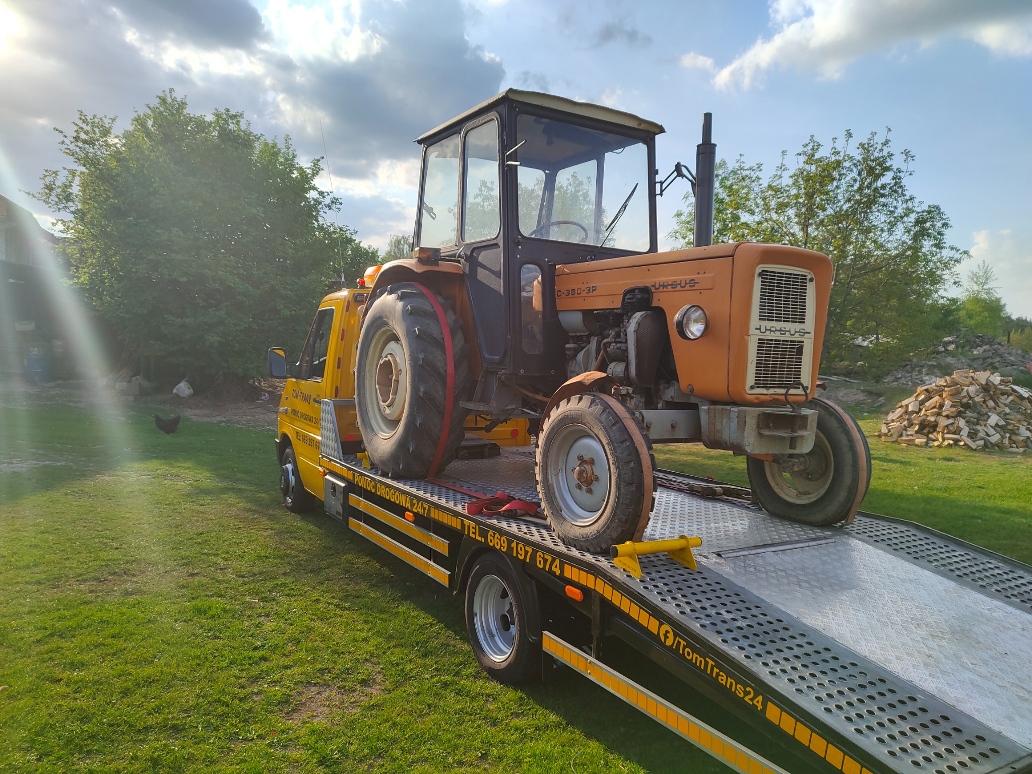 Żółta laweta z pomarańczowym traktorem Ursus C-360-3P na platformie, zabezpieczonym czerwonym pasem, na tle zielonego trawnika i błękitnego nieba z chmurami.