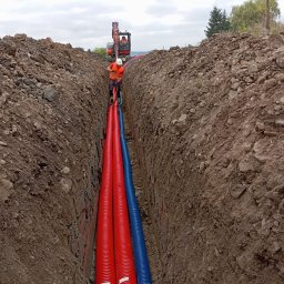 F.W NK-BUD KAZIMIERZ NIŻNIK - Wykopany rów z trzema rurami karbowanymi: dwiema czerwonymi i jedną niebieską, pracownik w kamizelce odblaskowej układa rury, a w tle stoi koparka.