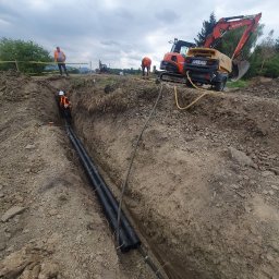 F.W NK-BUD KAZIMIERZ NIŻNIK - Głęboki wykop w ziemi z ułożonymi czarnymi rurami, pracownicy w odblaskowych kamizelkach i kaskach, pomarańczowa koparka Kubota z agregatem prądotwórczym w tle, zabezpieczenia wykopu taśmą.