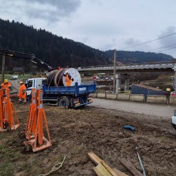 F.W NK-BUD KAZIMIERZ NIŻNIK - Prace instalacyjne przy budowie mostu: robotnicy w pomarańczowych kombinezonach rozładowują ciężkie zwoje kabli z ciężarówki za pomocą dźwigu, w tle konstrukcja mostu.