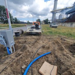 F.W NK-BUD KAZIMIERZ NIŻNIK - Szara skrzynka z przewodami i niebieską rurą na ziemi, samochód dostawczy z bębnami kablowymi w tle, robotnik w pomarańczowej kamizelce przy drodze, w oddali budynki przemysłowe.