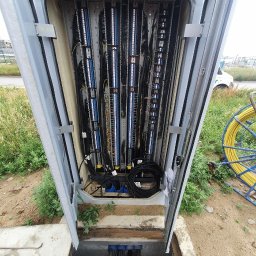 F.W NK-BUD KAZIMIERZ NIŻNIK - Otwarta skrzynka telekomunikacyjna z widocznymi kablami, złączami i panelami krosowniczymi, z boku widoczny bęben z kablem światłowodowym.