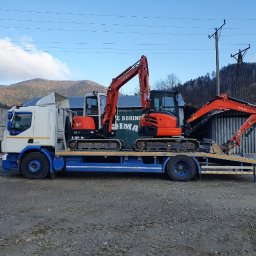 F.W NK-BUD KAZIMIERZ NIŻNIK - Ciężarówka z dwoma pomarańczowymi minikoparkami Kubota na platformie, w tle widoczne góry i betonowe bloki.