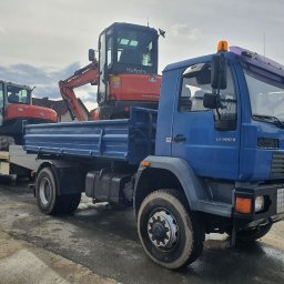 F.W NK-BUD KAZIMIERZ NIŻNIK - Niebieska ciężarówka MAN z przyczepą, transportująca dwie pomarańczowe mini koparki Kubota na placu budowy, widoczne góry w tle.