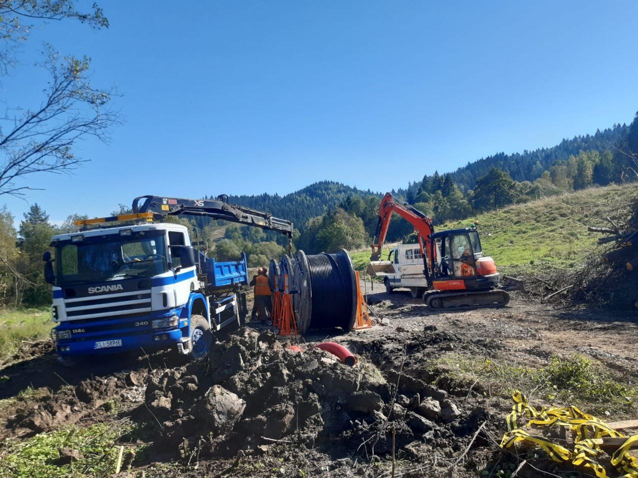 Rozładunek bębna z kablem telekomunikacyjnym za pomocą dźwigu samochodowego Scania oraz koparki Kubota, w tle zalesione wzgórza.