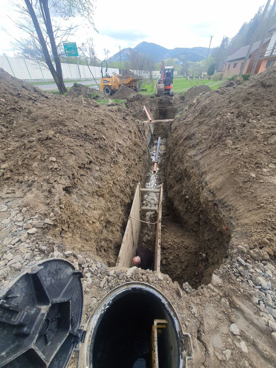 Głęboki wykop z drewnianym szalunkiem, wewnątrz którego pracuje osoba przy instalacji rur. W tle maszyny budowlane i krajobraz zalesionych wzgórz. Otwarta studzienka kanalizacyjna na pierwszym planie.