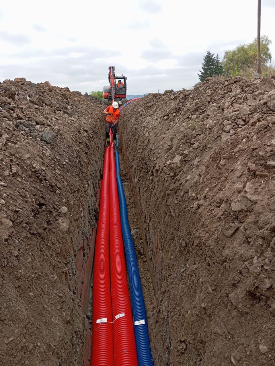 Wykopany rów z trzema rurami karbowanymi: dwiema czerwonymi i jedną niebieską, pracownik w kamizelce odblaskowej układa rury, a w tle stoi koparka.