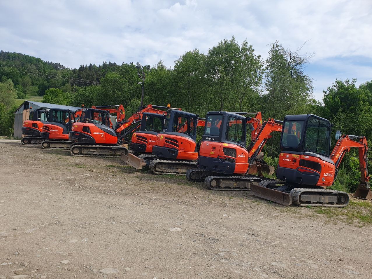 Rząd pomarańczowych minikoparek Kubota zaparkowanych na żwirowym terenie, zalesione wzgórze w tle.