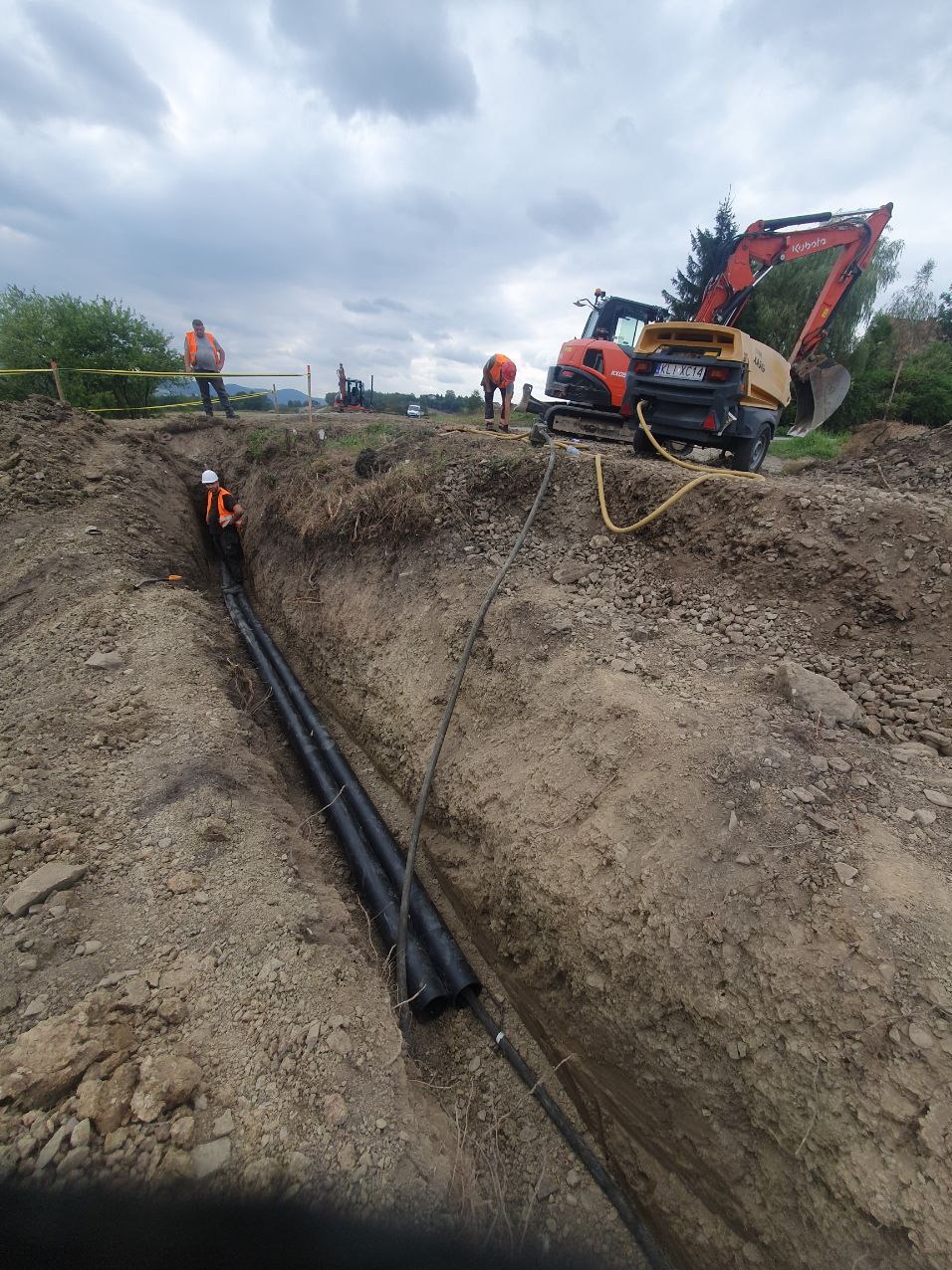 Głęboki wykop w ziemi z ułożonymi czarnymi rurami, pracownicy w odblaskowych kamizelkach i kaskach, pomarańczowa koparka Kubota z agregatem prądotwórczym w tle, zabezpieczenia wykopu taśmą.