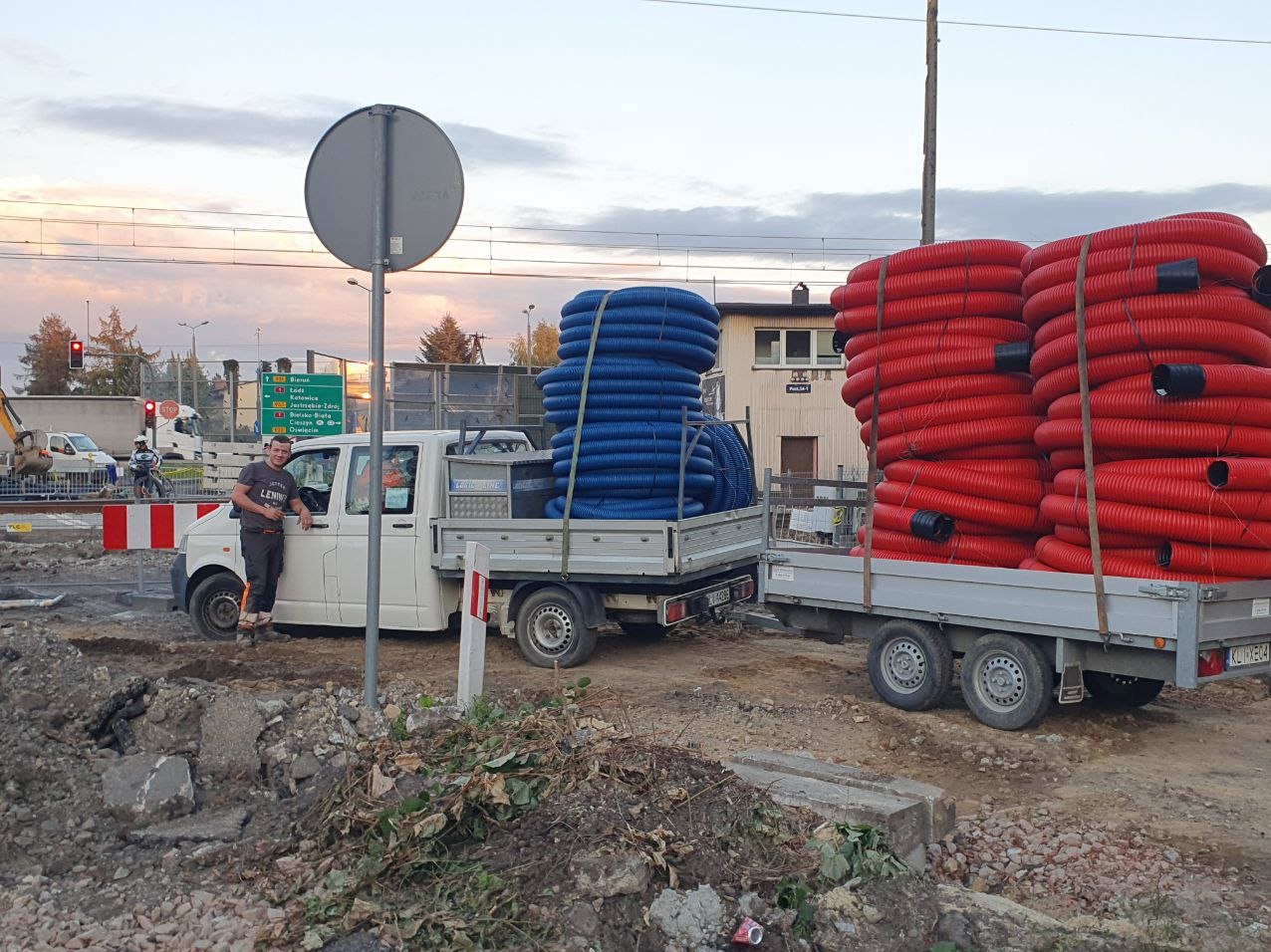 Pracownik opiera się o białego dostawczaka z niebieskimi rurami karbowanymi na pace, a na przyczepce czerwone rury karbowane; w tle rozkopana ulica i budynek.