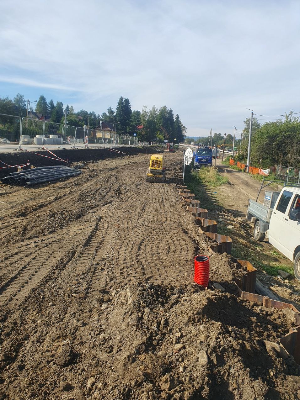 Przygotowanie terenu pod budowę: wyrównana ziemia, widoczne ślady opon, mała koparka, rury, metalowe zabezpieczenia wykopu i samochody w tle.