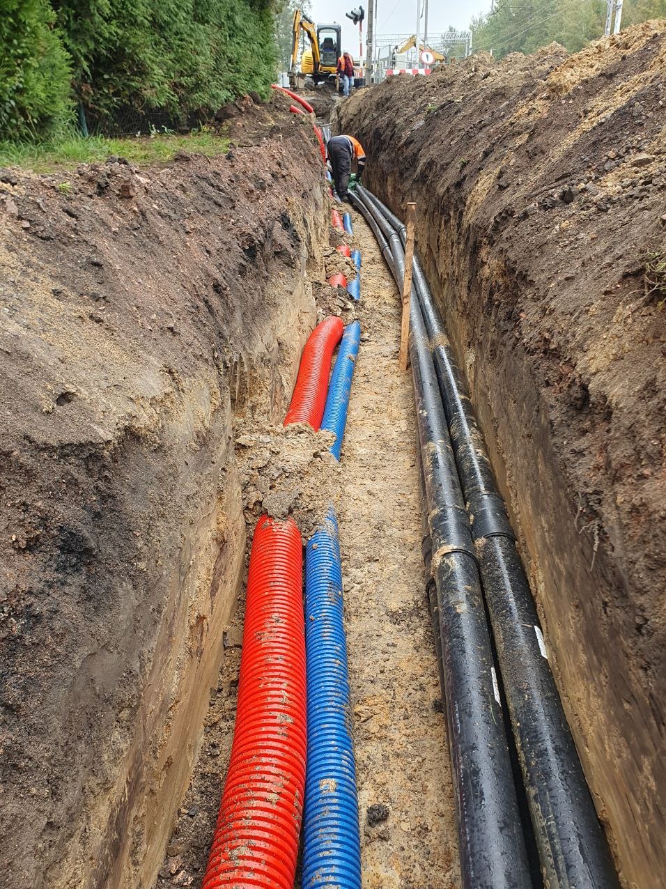 Wykop w ziemi z ułożonymi rurami w kolorach czerwonym, niebieskim i czarnym. W tle widoczny pracownik w kombinezonie i koparka.