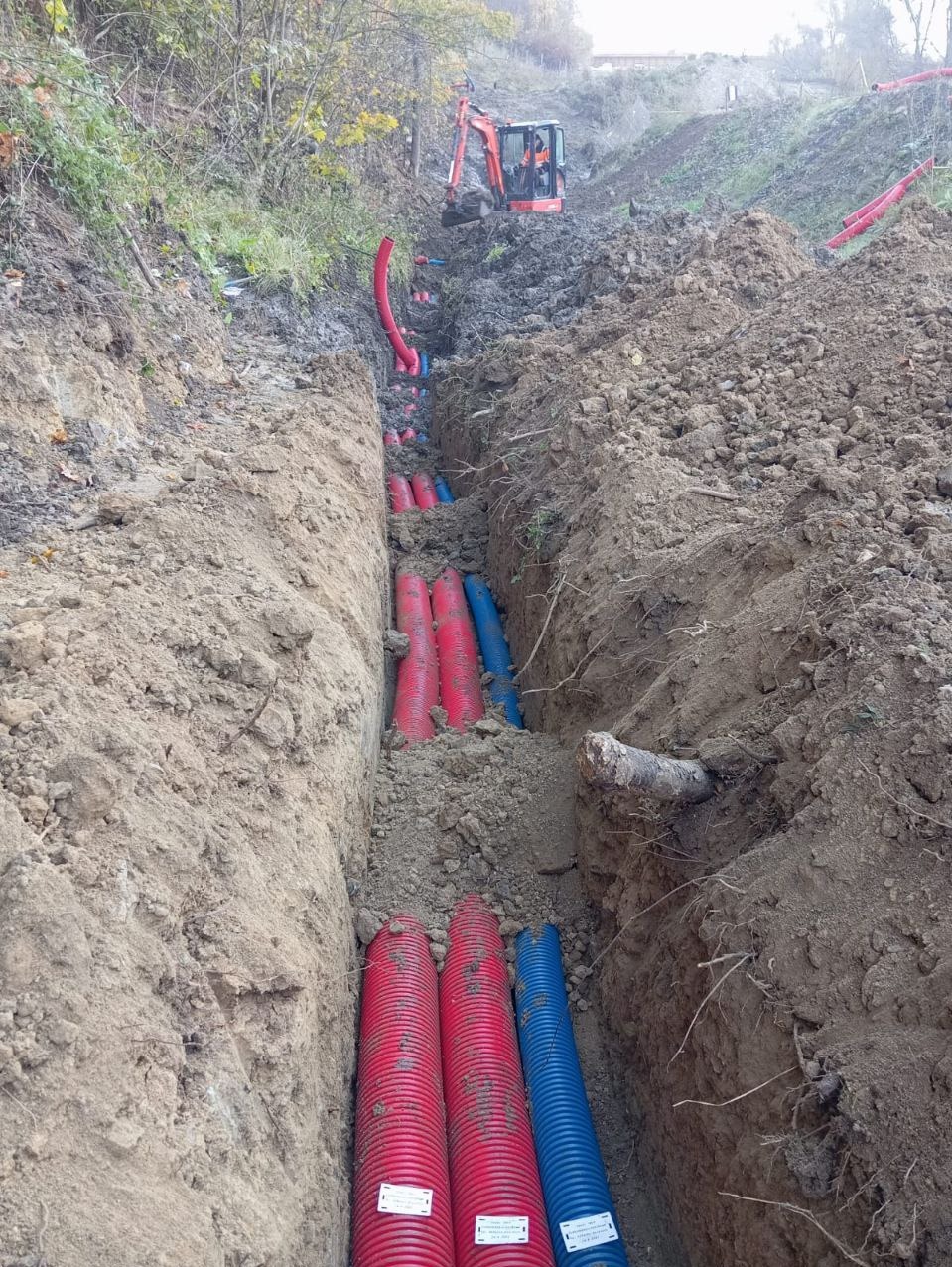 Głęboki wykop z ułożonymi rurami osłonowymi w kolorach czerwonym i niebieskim, z widoczną koparką na zboczu w tle.