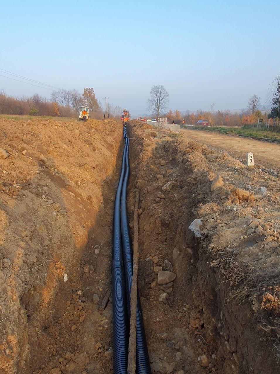 Dwa czarne, karbowane przewody w głębokim wykopie ziemnym; w tle widoczny sprzęt budowlany i drzewa.