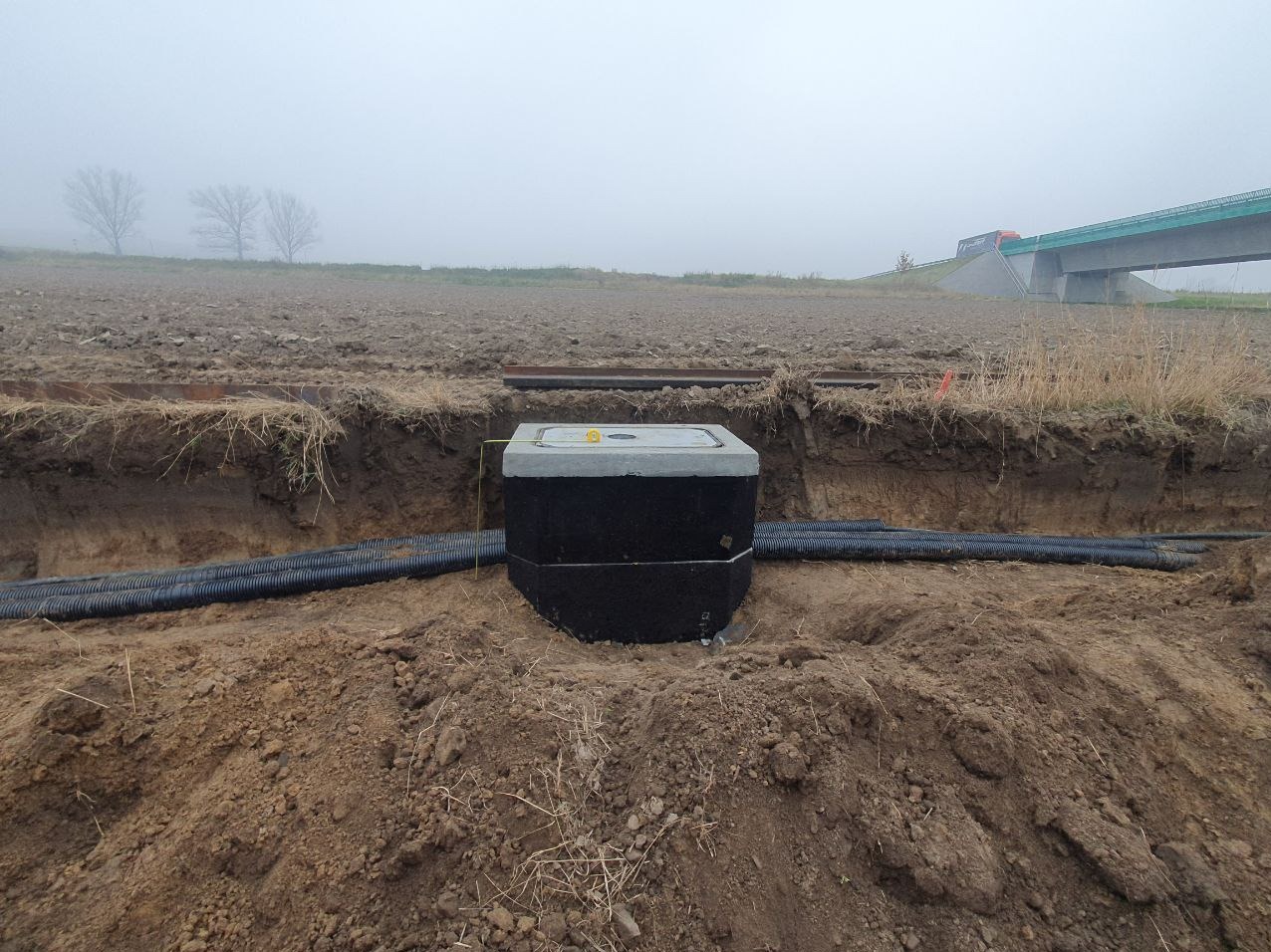 Betonowa studzienka z szarym wiekiem i żółtym uchwytem, umieszczona w wykopie ziemnym. Do studzienki dochodzą rury karbowane z tworzywa sztucznego. W tle widoczny most i zamglone pole.