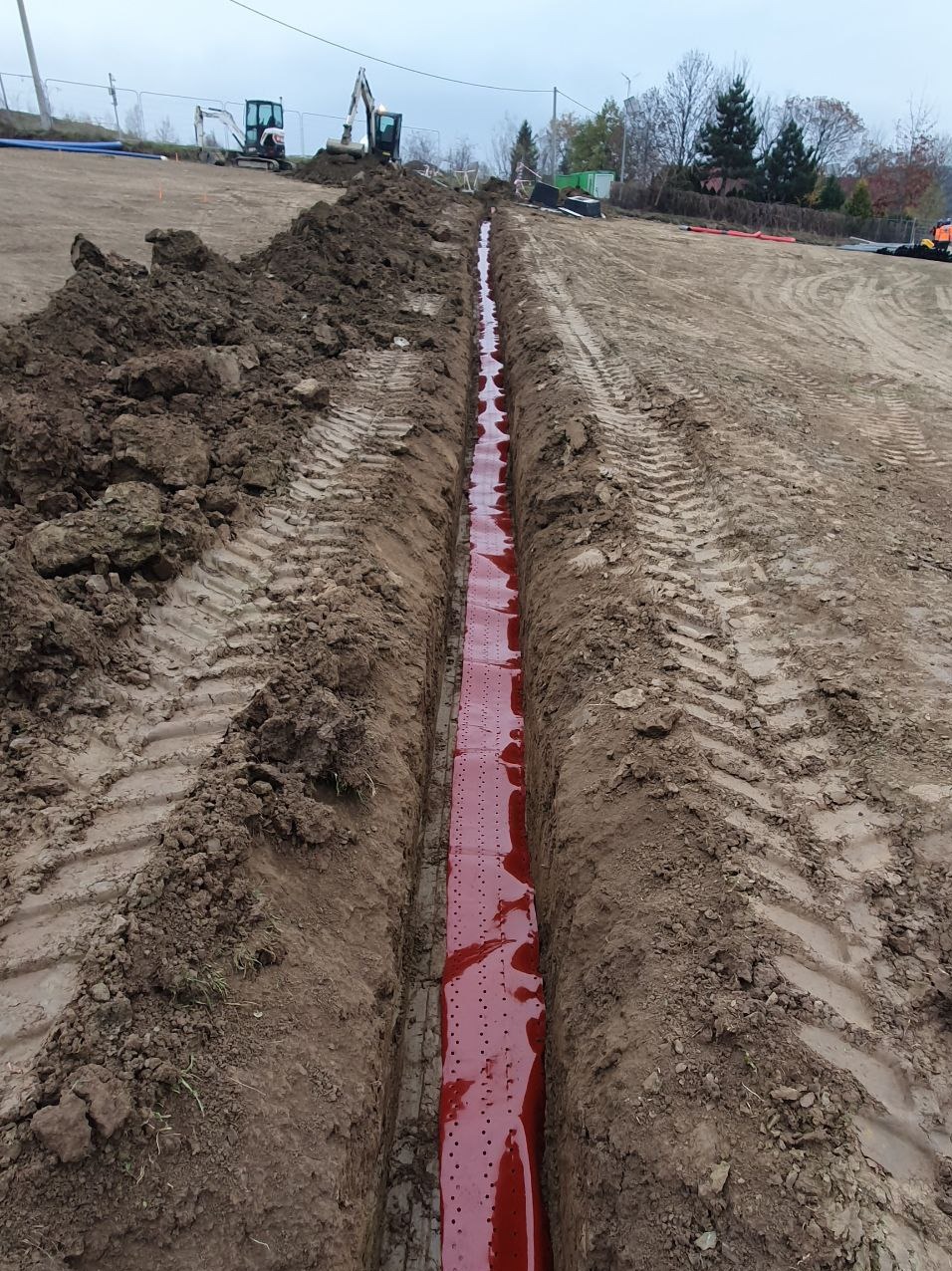 Długi, wąski wykop w ziemi z widoczną czerwoną perforowaną folią ochronną na dnie, ślady opon koparki po obu stronach, dwie koparki w tle, pochmurne niebo.