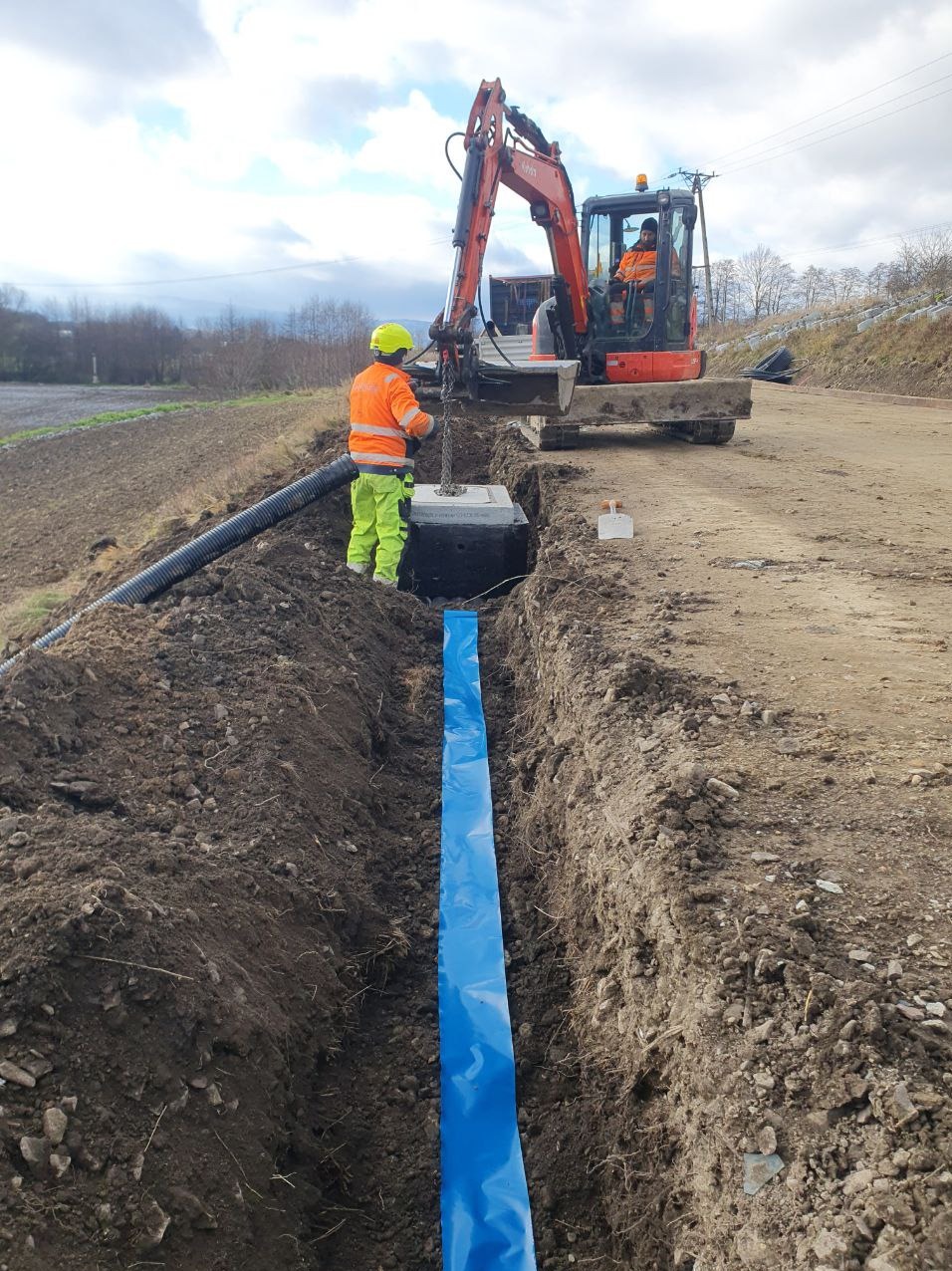 Pracownik w odblaskowej odzieży i kasku kieruje betonowy element za pomocą łańcucha podczepionego do koparki w wykopie wyłożonym niebieską folią. W tle widoczny drugi pracownik w koparce i krajobraz.