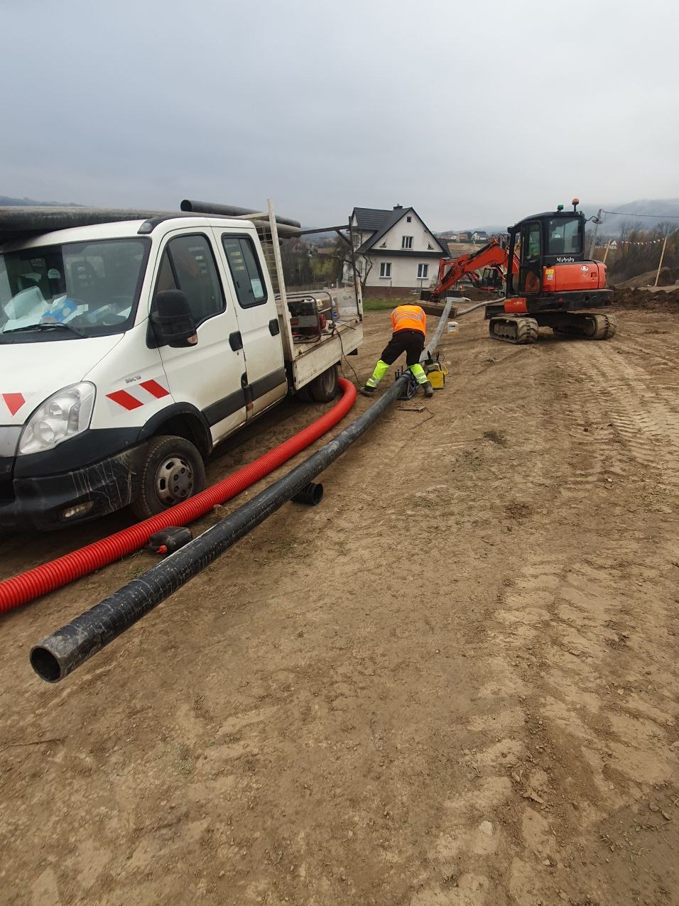 Pracownik w kamizelce odblaskowej łączy rury na placu budowy, w tle biały samochód dostawczy, koparka i dom w oddali.