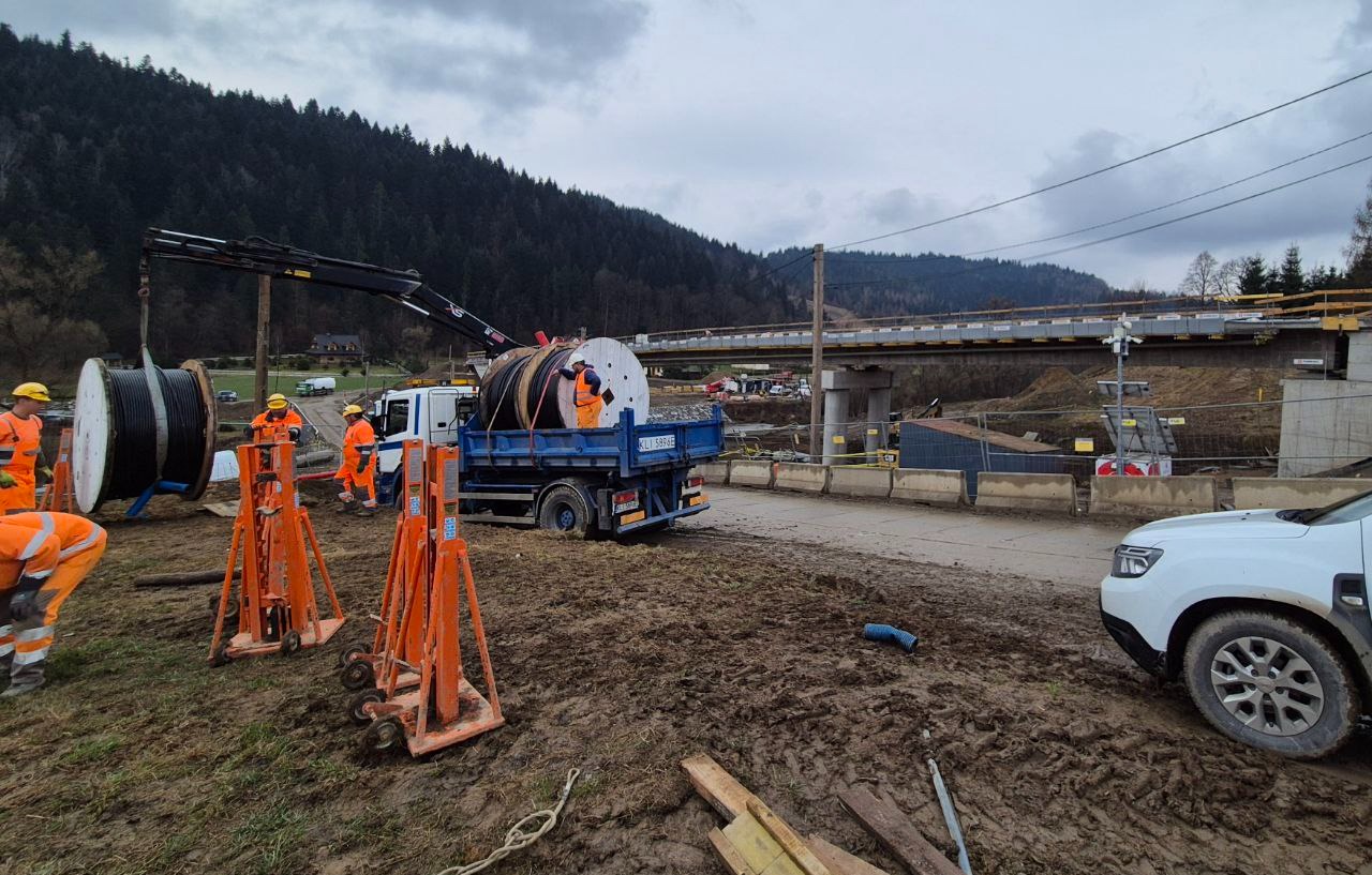 Prace instalacyjne przy budowie mostu: robotnicy w pomarańczowych kombinezonach rozładowują ciężkie zwoje kabli z ciężarówki za pomocą dźwigu, w tle konstrukcja mostu.