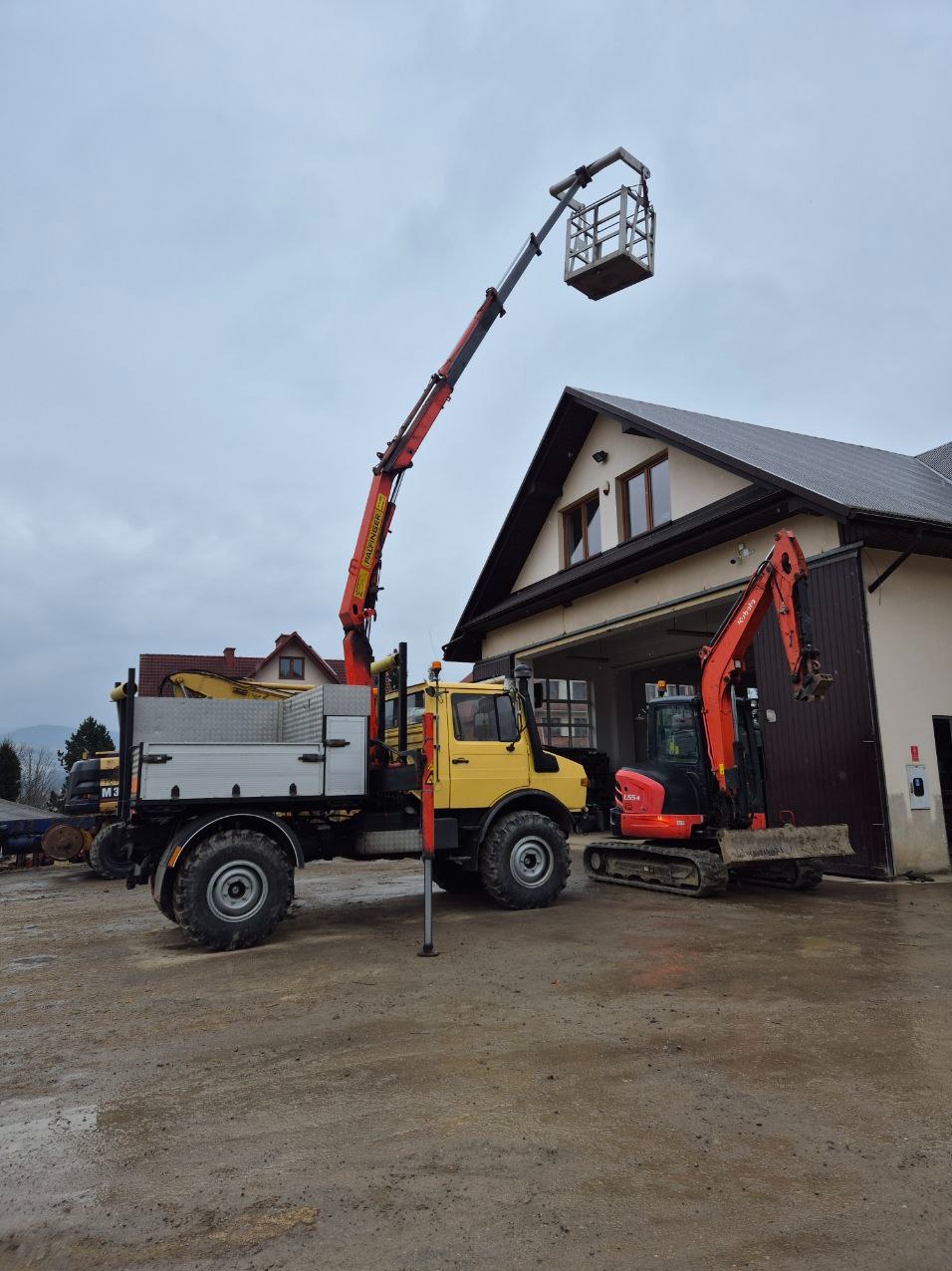 Żółty Unimog z podnośnikiem koszowym Palfinger oraz pomarańczowa mini koparka Kubota na placu budowy przed budynkiem z bramą garażową.