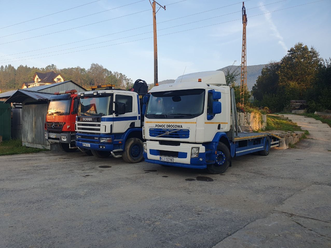 Trzy samochody pomocy drogowej różnych marek (Mercedes, Scania, Volvo) zaparkowane obok siebie na betonowym placu, w tle budynki i zalesione wzgórza.