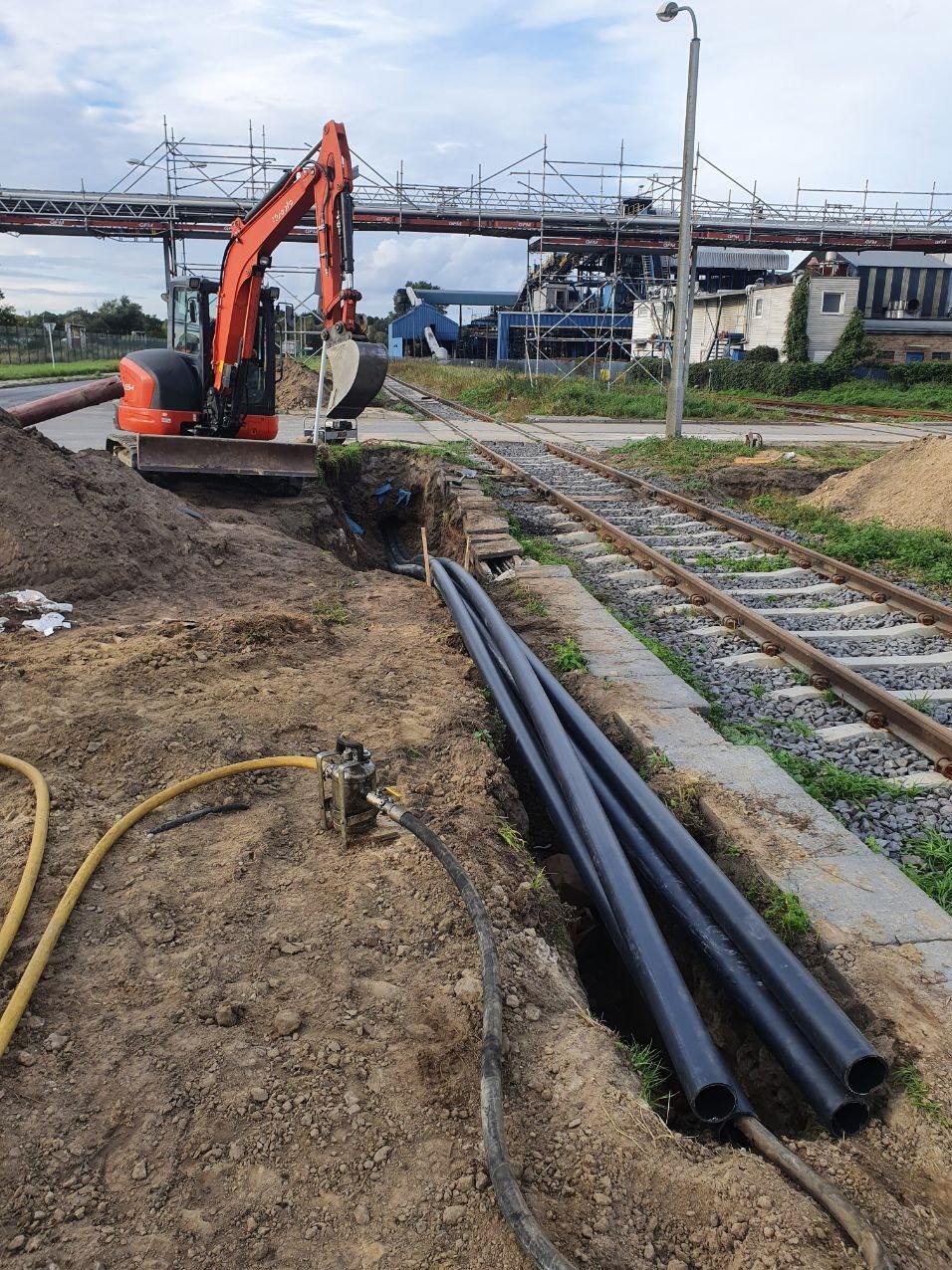 Pomarańczowa mini koparka Kubota przygotowuje wykop pod rury w pobliżu torów kolejowych, widoczny fragment instalacji z żółtym wężem i cztery czarne rury ułożone w wykopie, w tle zabudowania...