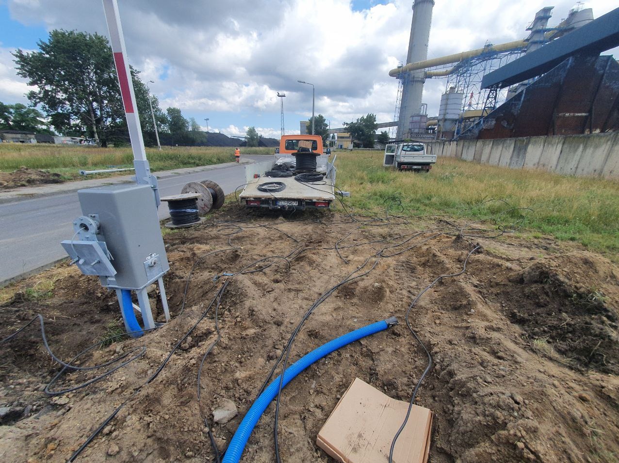 Szara skrzynka z przewodami i niebieską rurą na ziemi, samochód dostawczy z bębnami kablowymi w tle, robotnik w pomarańczowej kamizelce przy drodze, w oddali budynki przemysłowe.
